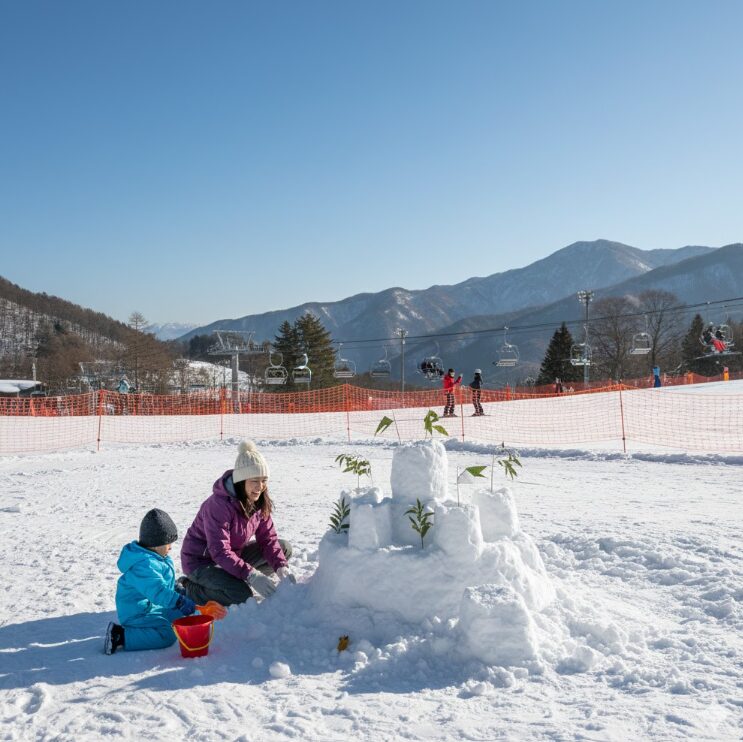 スキー・スノーボードエリアとは明確にネットで区切られた、平坦で広い雪遊び広場。日本人の親子がスコップやバケツを使って雪の城を作っている。遠くにリフトが見えるが、手前の広場はゆったりとした時間が流れている。
